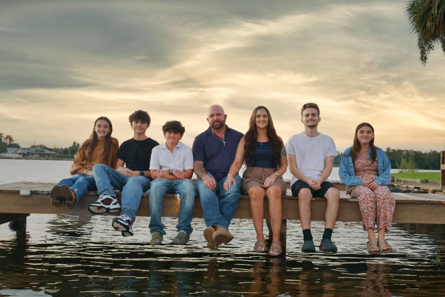 Family of seven sits in a row on a wooden dock over the water at sunset for a warm, relaxed group portrait.