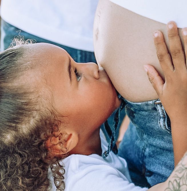 Little girl with curly hair tenderly kisses her pregnant mother's baby bump in a close-up maternity portrait.