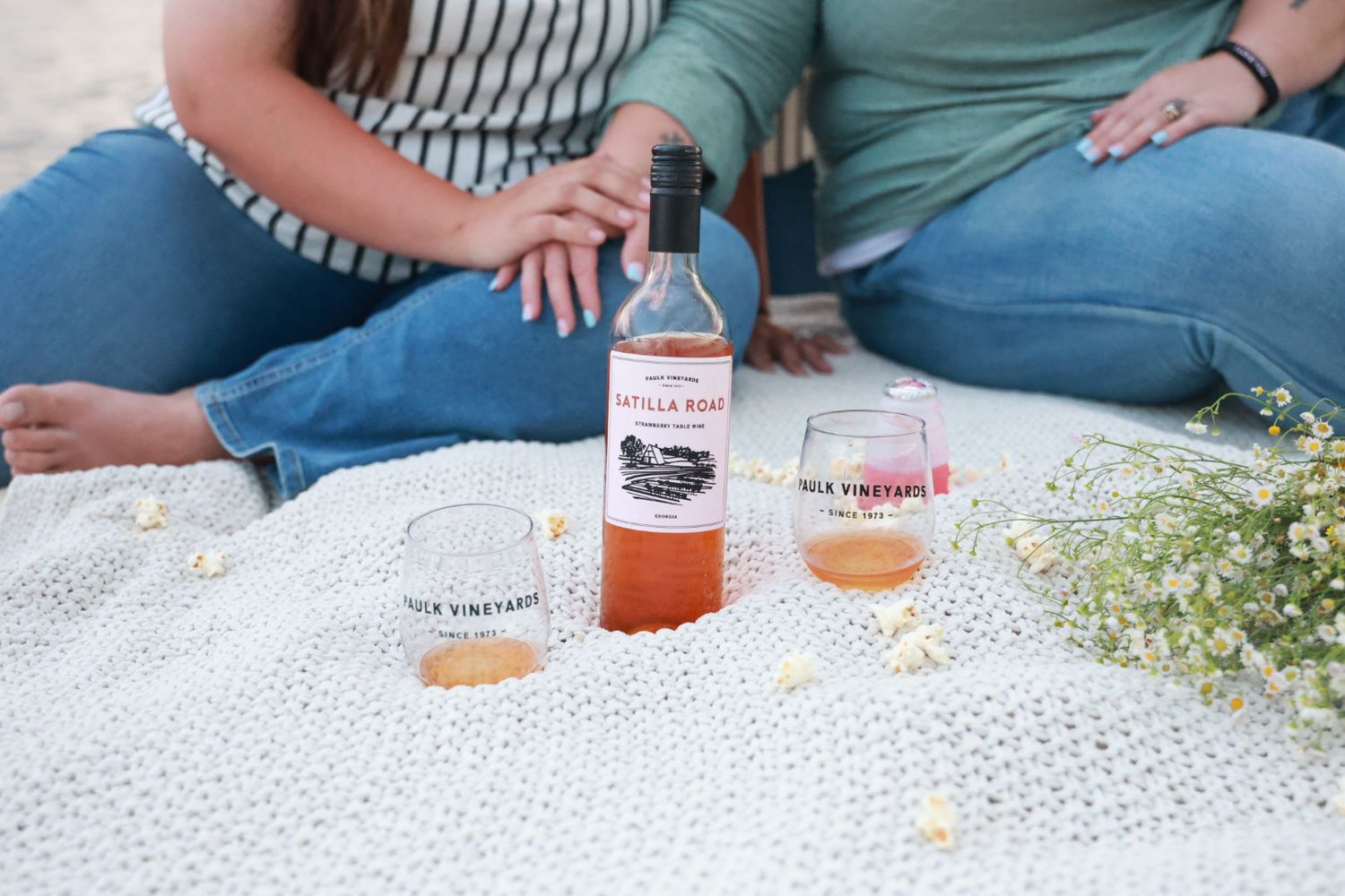 Couple relaxing on a white blanket with a bottle of rosé wine, two glasses, and white wildflowers during a lakeside picnic.
