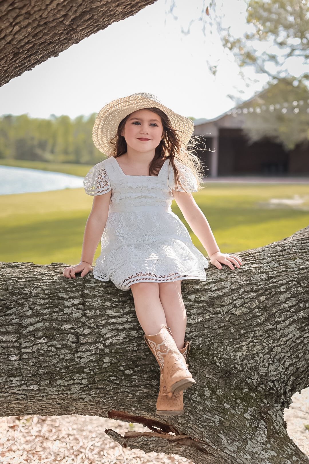 Smiling little girl in a white dress, sun hat, and cowboy boots perches on a thick oak branch with a pond in the background.