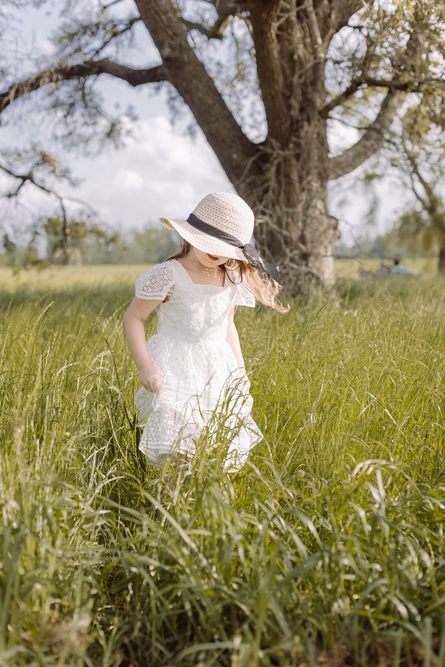 Little girl in a white dress and straw sun hat walking through a tall green meadow with a big oak tree behind her.