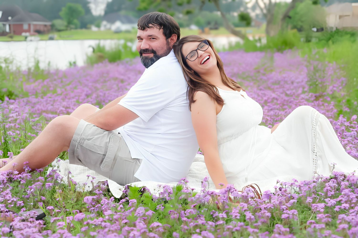 A bearded man and a laughing woman in glasses sit back to back in a blooming field of purple wildflowers.