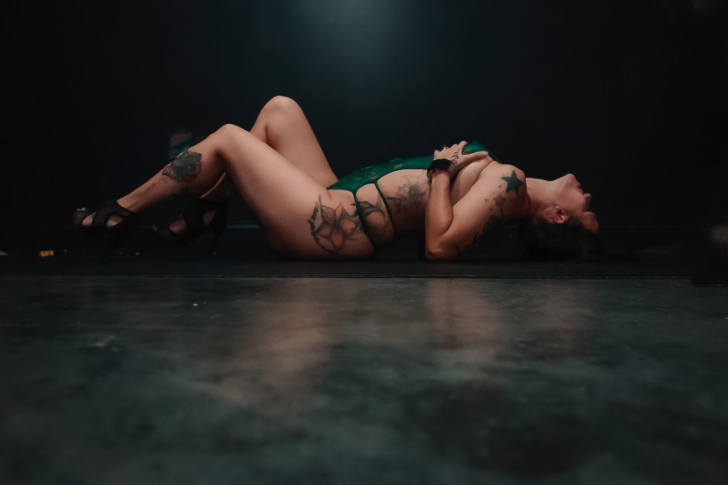 Tattooed woman in a green bodysuit and heels reclines on a reflective dark floor in a moody boudoir photography setting.