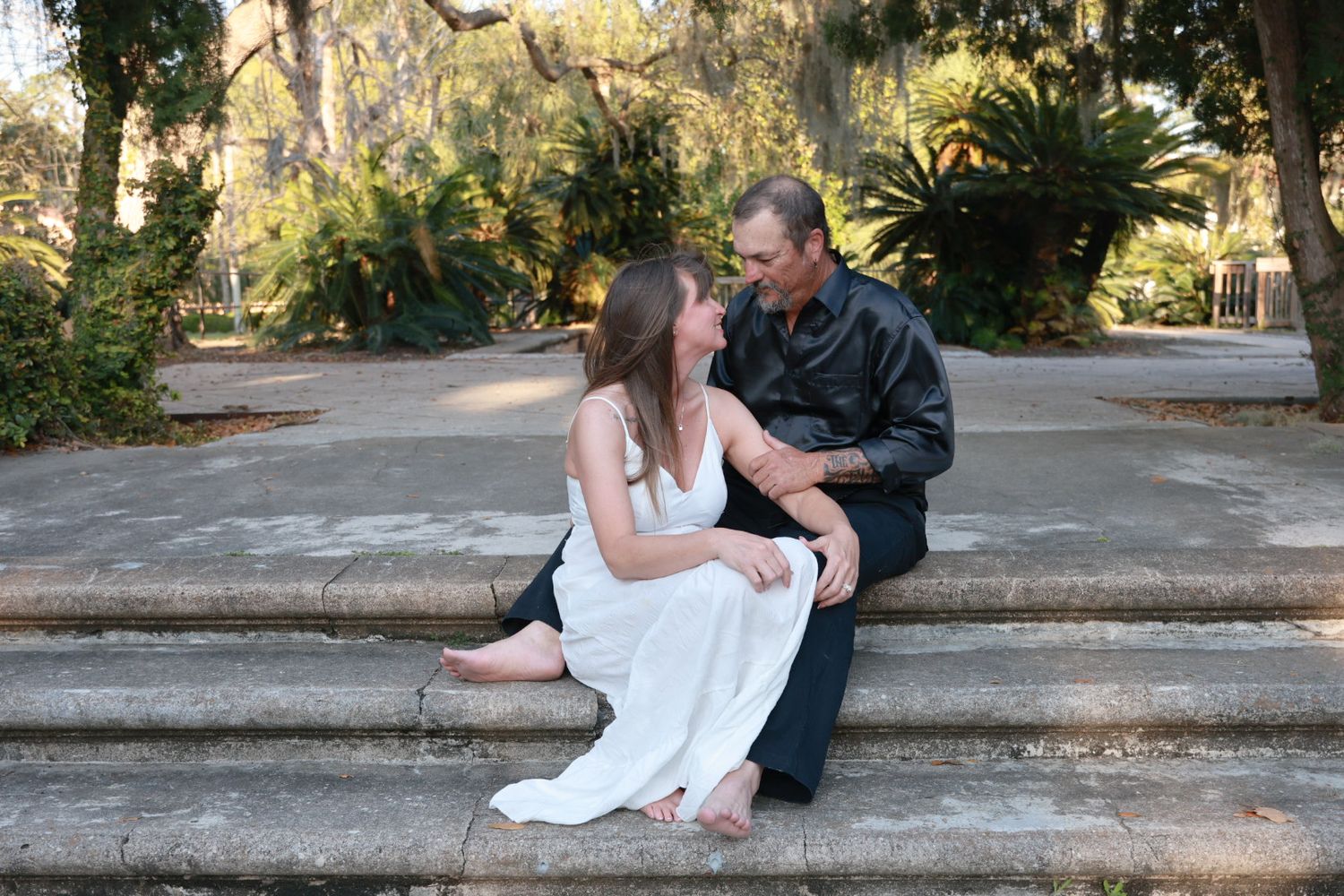 Couple sit close together on wide stone steps surrounded by lush greenery and trees in a garden.