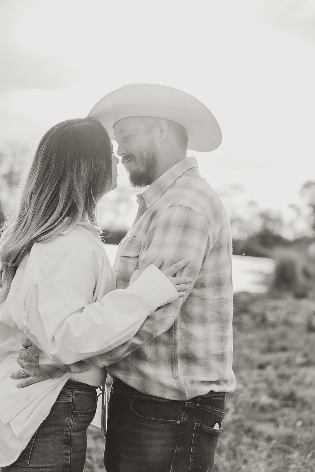 Black and white photo of a couple in cowboy hats and plaid embracing with noses touching in a sunlit field.
