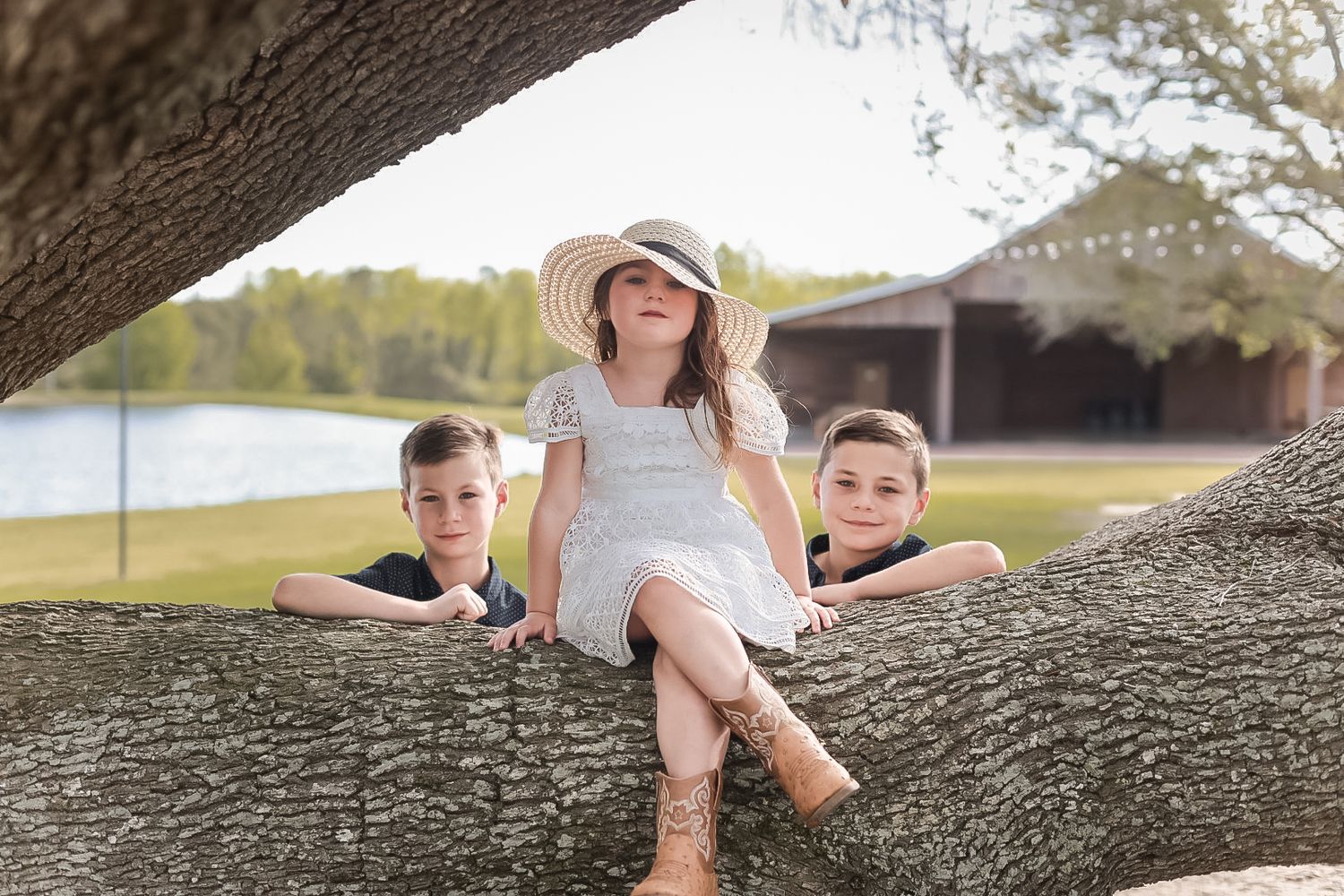 Young girl in a white dress, sun hat, and cowboy boots sits on a tree limb with two boys leaning beside her near a lake.