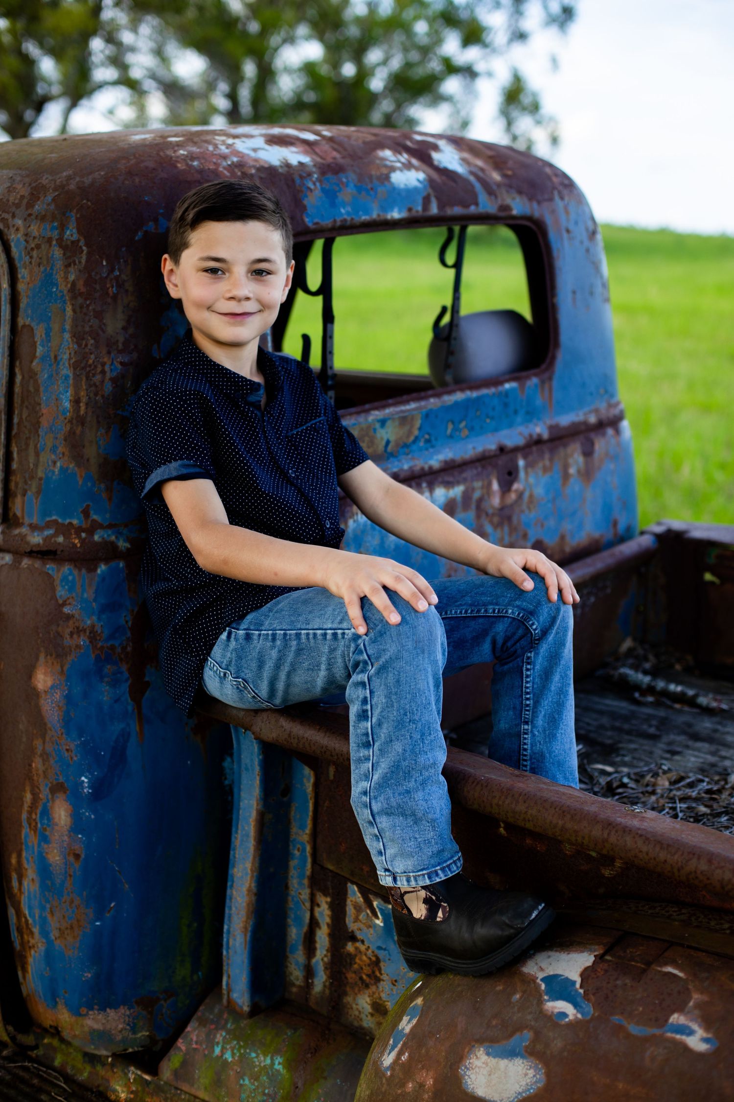 Smiling boy in a navy shirt and jeans sits on the tailgate of a rusty vintage blue truck in a grassy field.