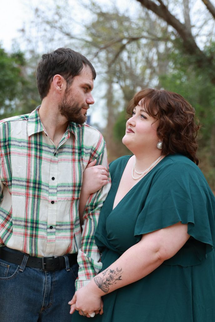 Bearded man in a plaid shirt gazes at his partner in an emerald green dress as they stand arm in arm among trees.