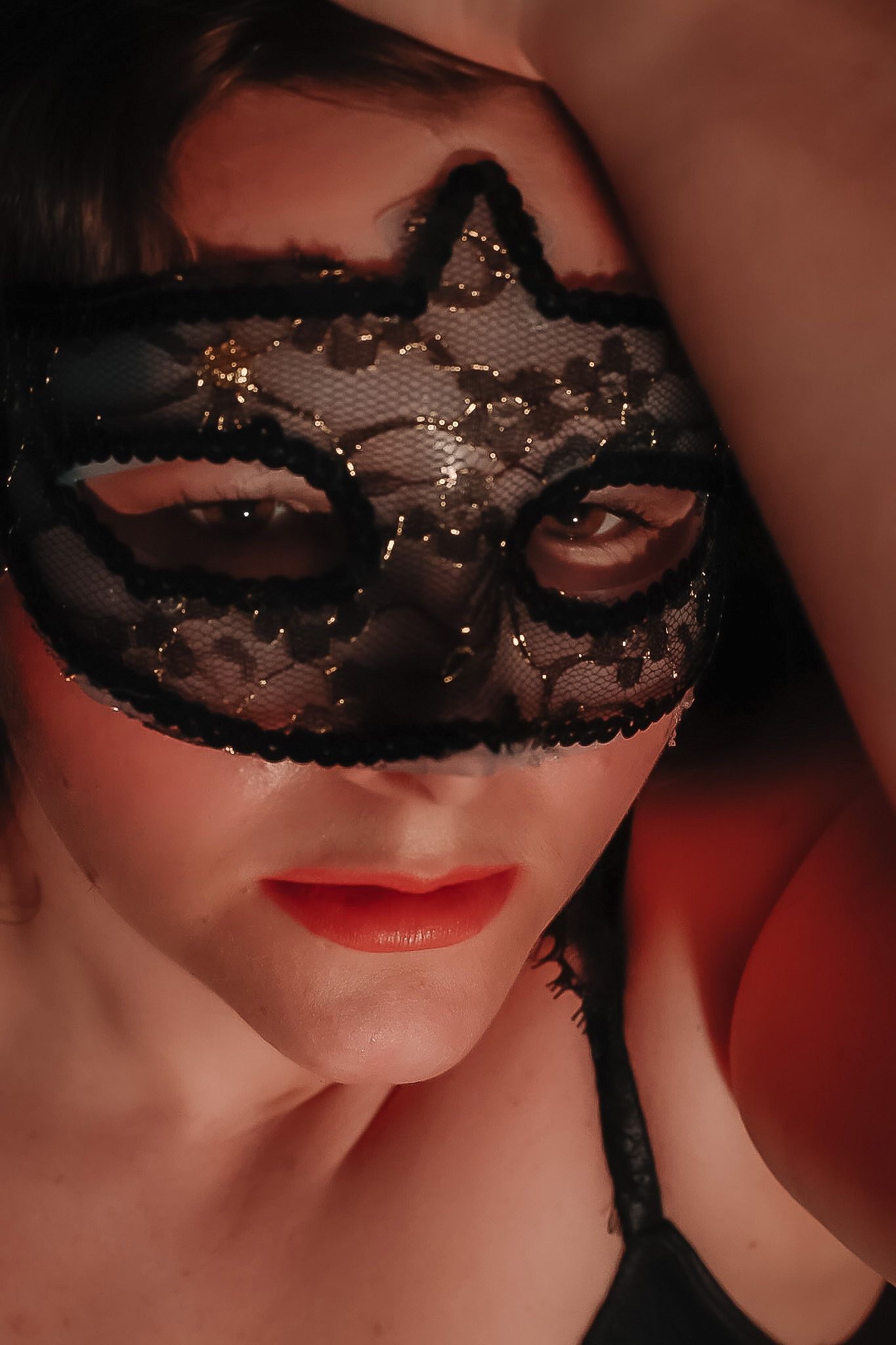 Close-up portrait of a woman in a black lace masquerade mask with red lipstick, her arm raised above her head.