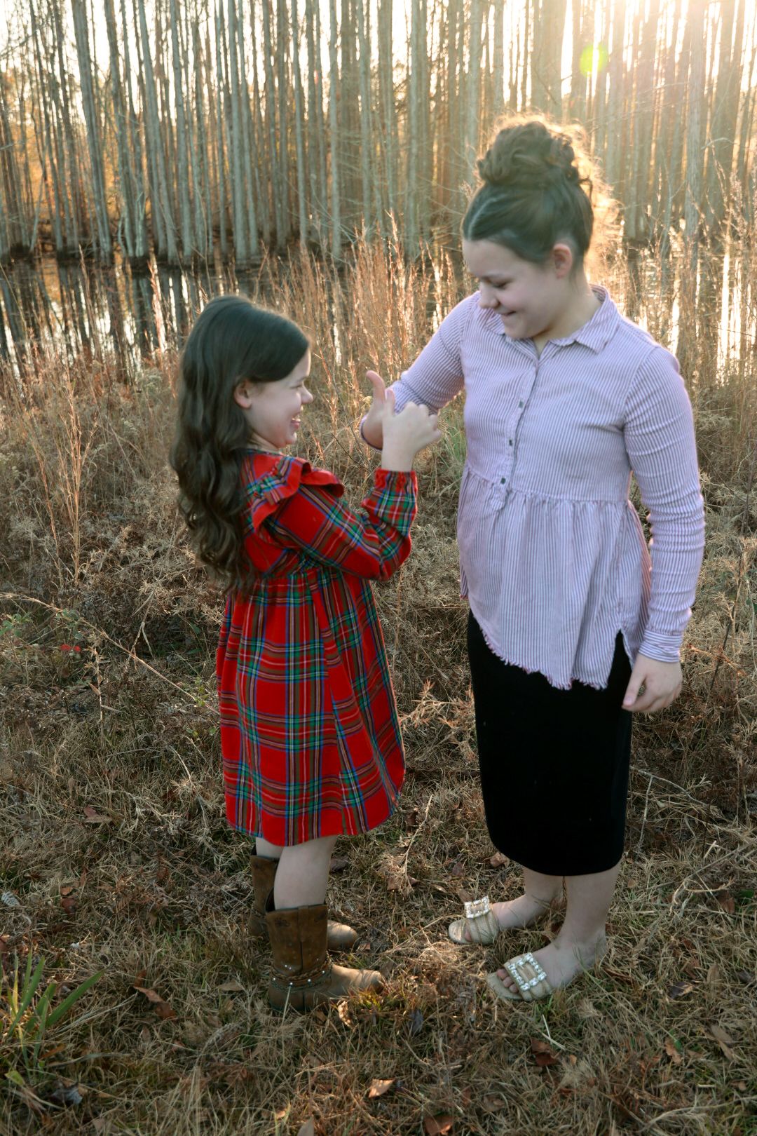 Two girls hold hands in a grassy field by a pond at sunset, one in a red plaid dress and one in a lavender blouse.