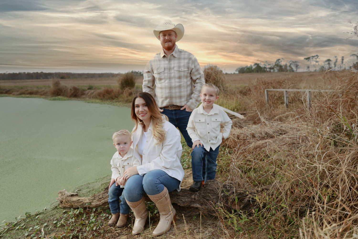 A cowboy-hat-wearing family of four poses by a green pond at sunset, with a mother and two young sons seated in front of dad.