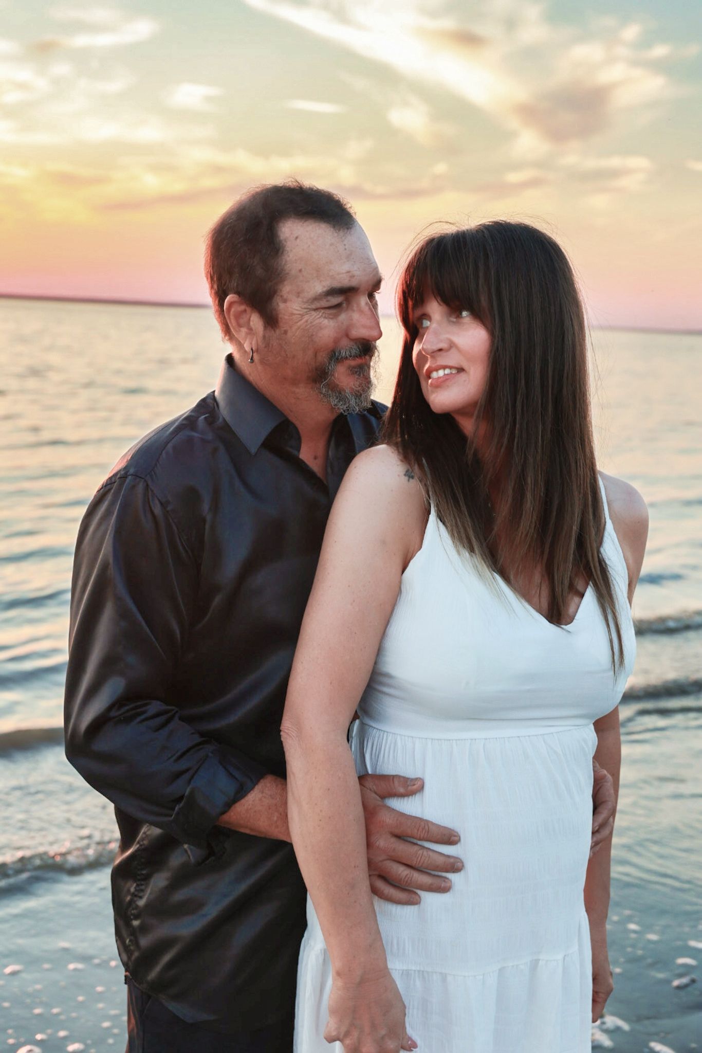 A smiling man in a dark shirt embraces a woman in a white dress from behind on the beach as the sunset glows over the water.