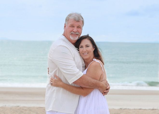 Smiling couple in white outfits hug on a sandy beach with gentle ocean waves in the background.
