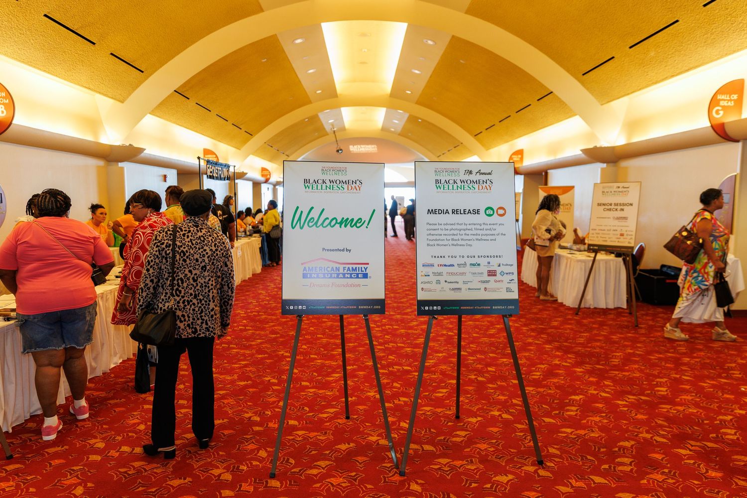 Welcome signs on easels greet attendees in a hotel conference area with red carpeting and warm lighting.