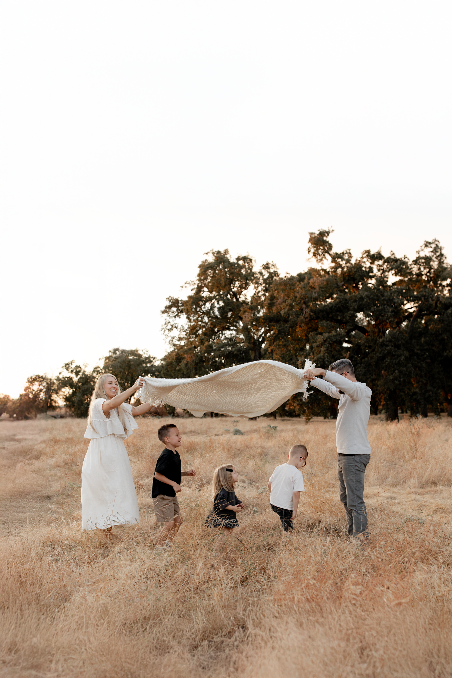 Family plays together in a golden field at sunset as they wave a white blanket in the air.
