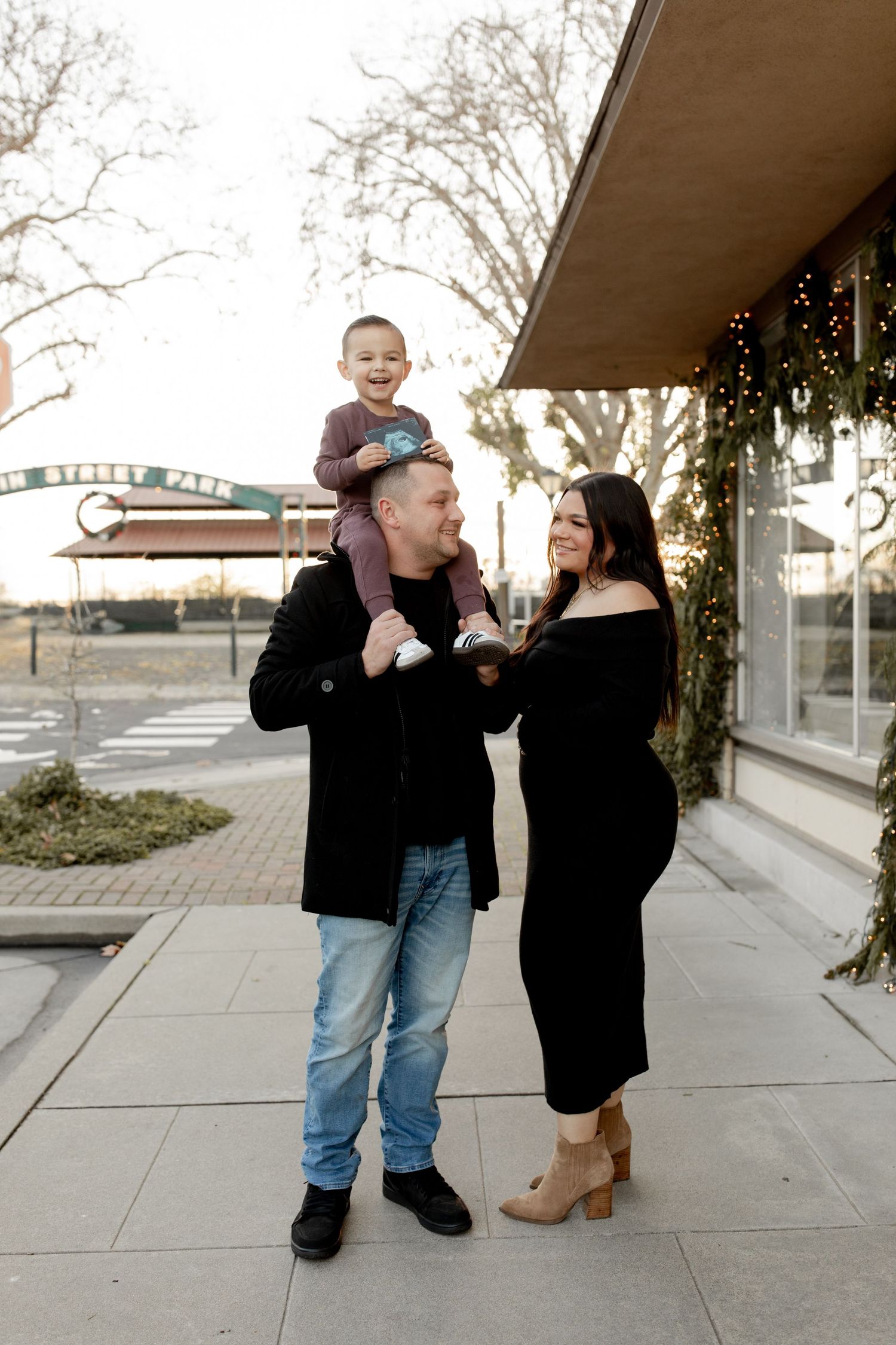A happy family poses outdoors, dad holds toddler on shoulders beside pregnant mom in black dress.