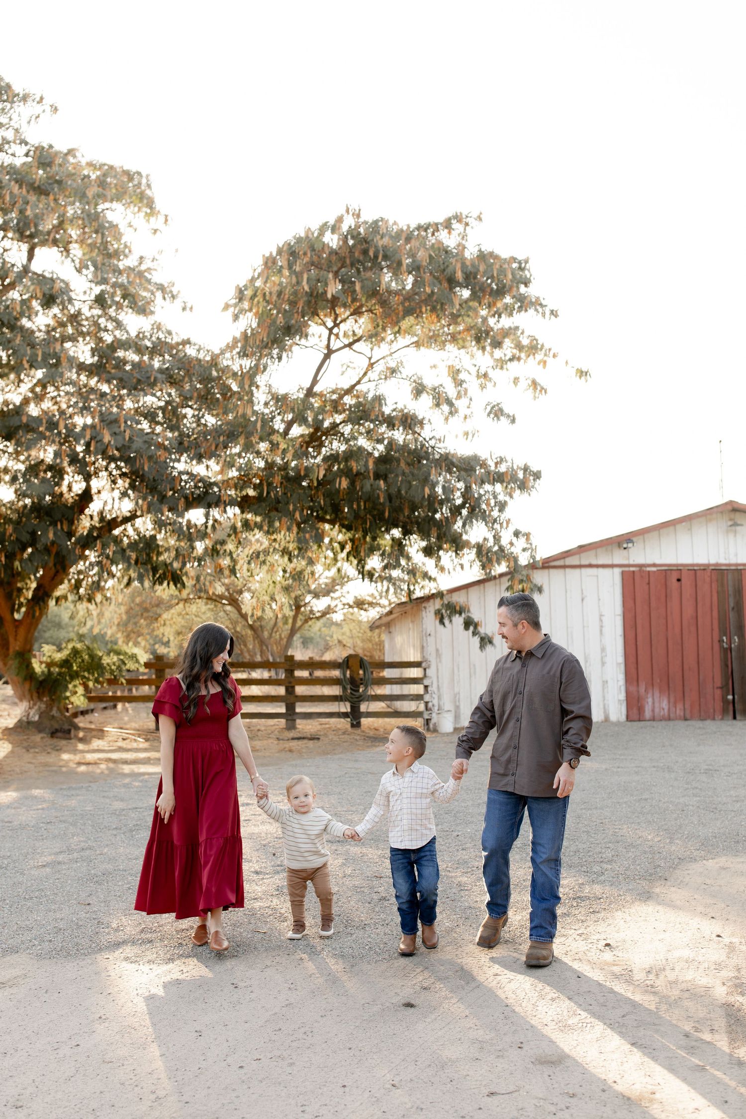 A family holds hands and walks together on a dirt path near a barn and tree during golden hour.