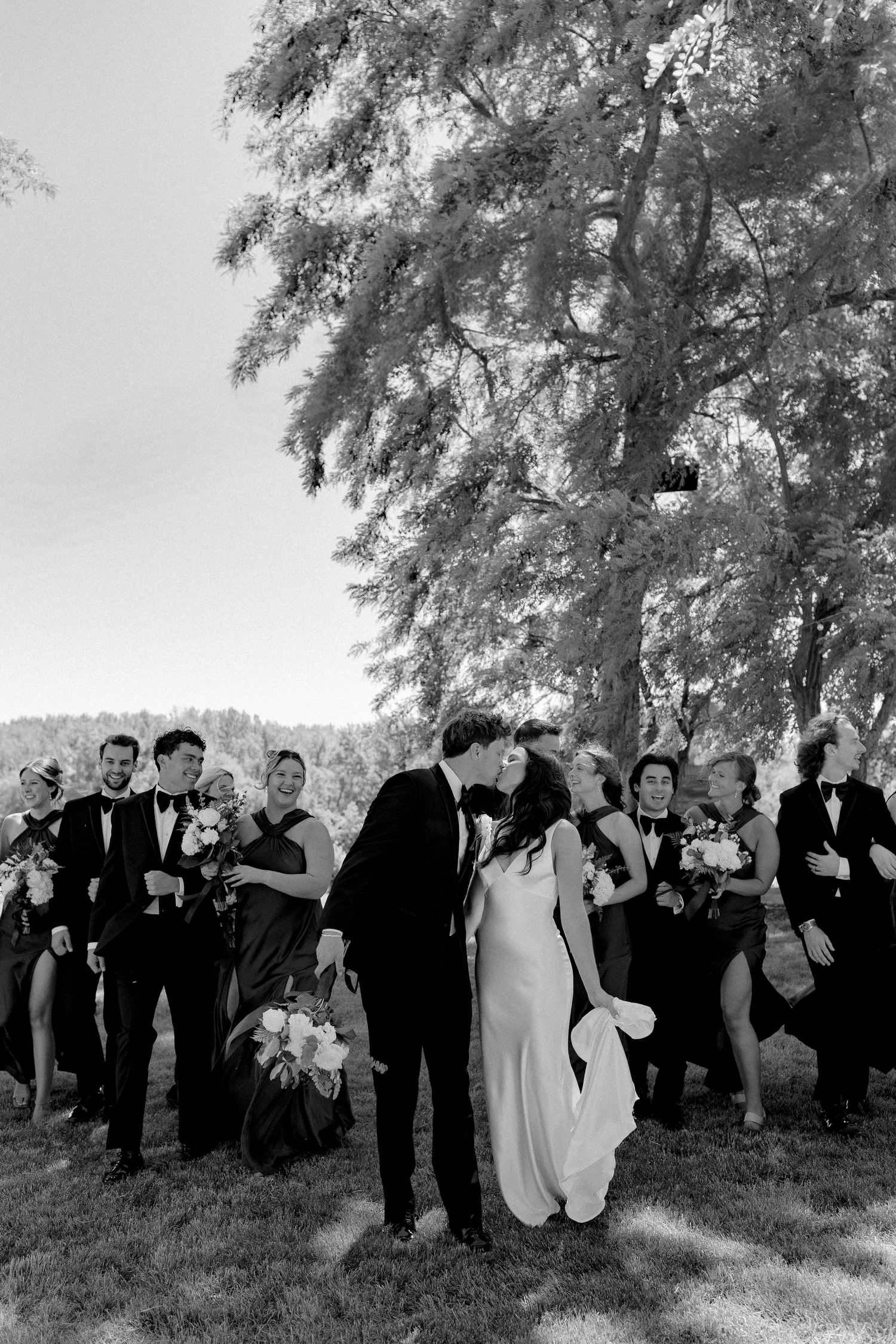 Black and white wedding photo of couple kissing surrounded by bridal party under a large tree outdoors.