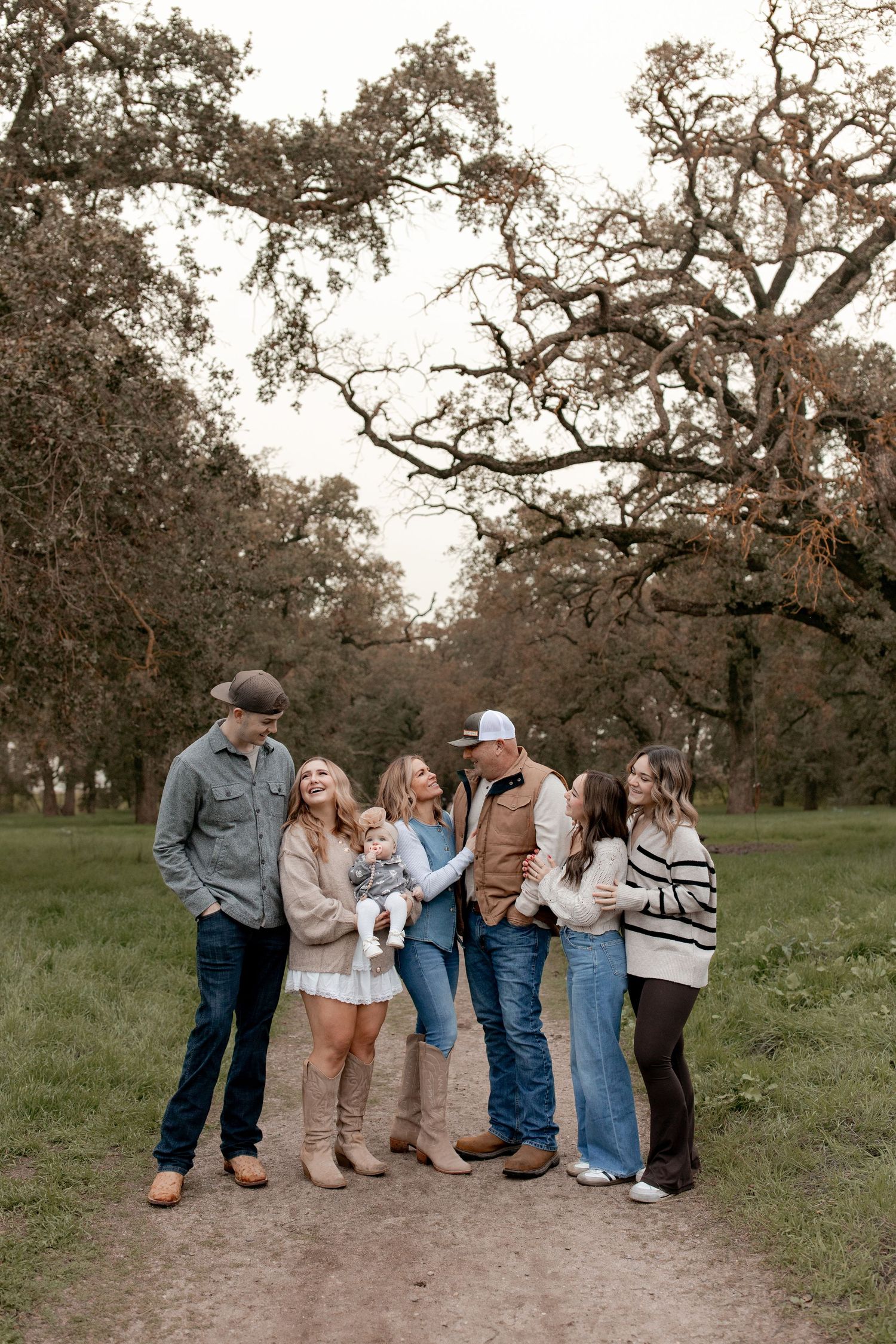 A group of people stand together in casual attire against a backdrop of oak trees during a golden hour photoshoot.