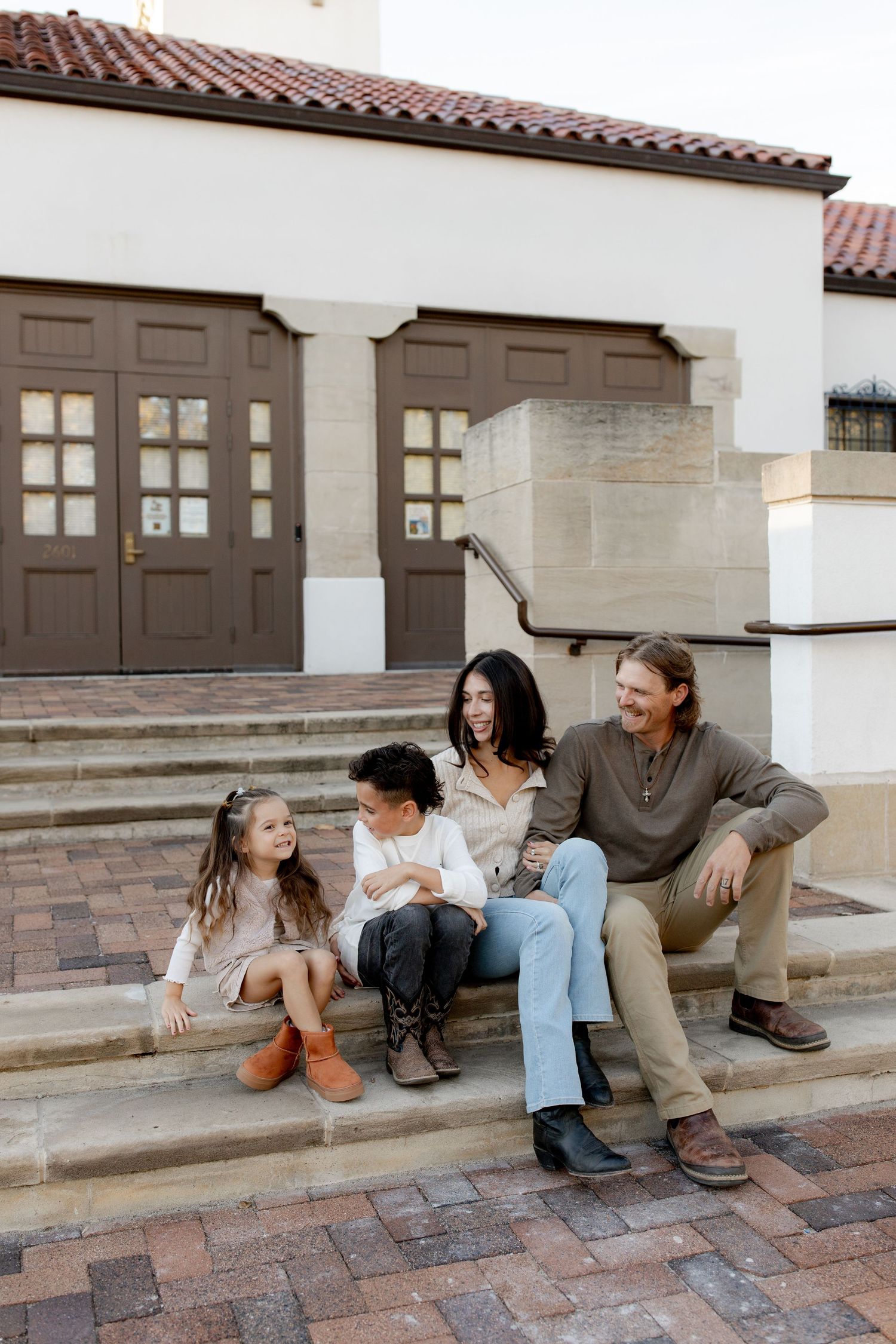 Family of four sitting on steps outside a Spanish-style building, smiling and enjoying a candid moment together.