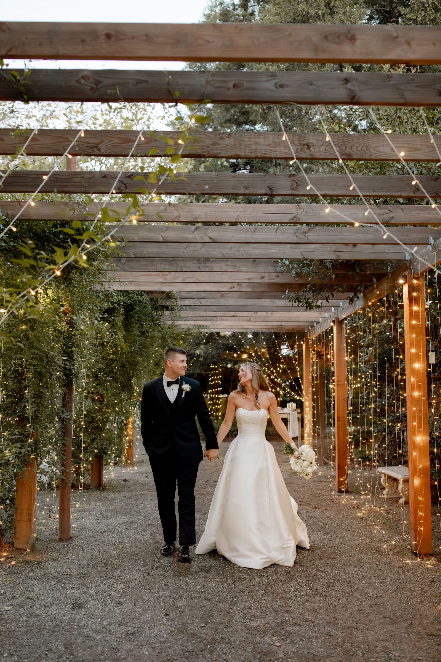 Bride and groom walk hand-in-hand under a wooden pergola adorned with fairy lights and greenery at their wedding.