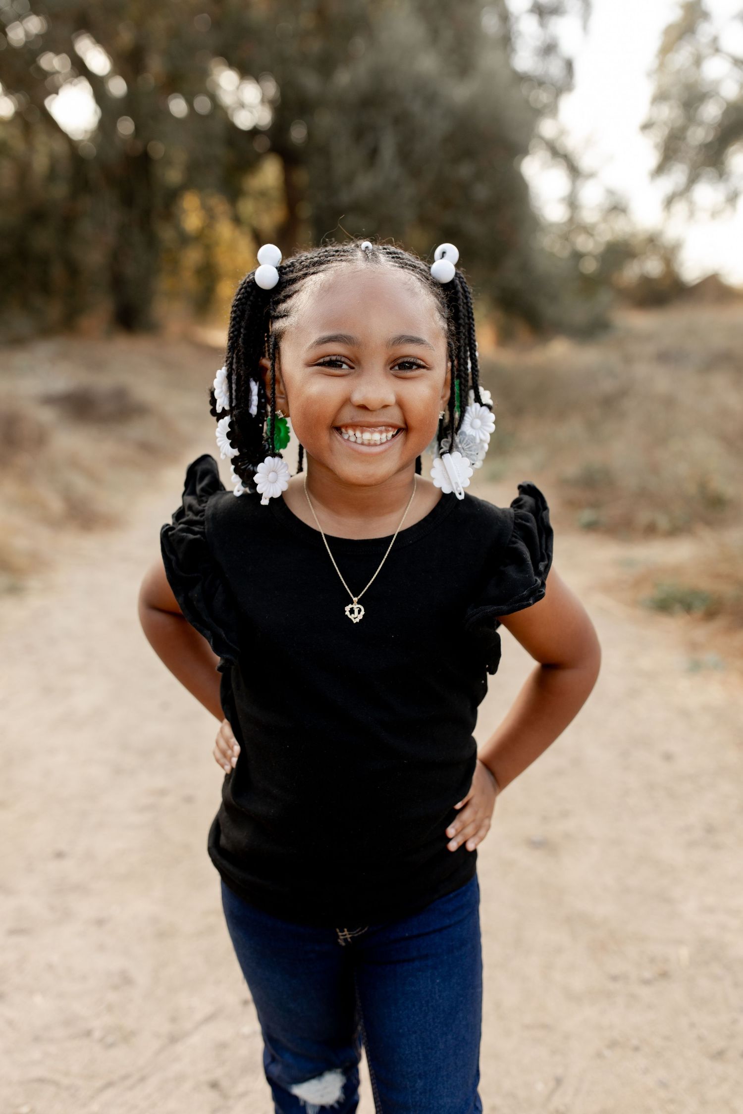 Child in a black ruffle sleeve top and jeans stands on a dirt path with hair accessories, smiling outdoors.