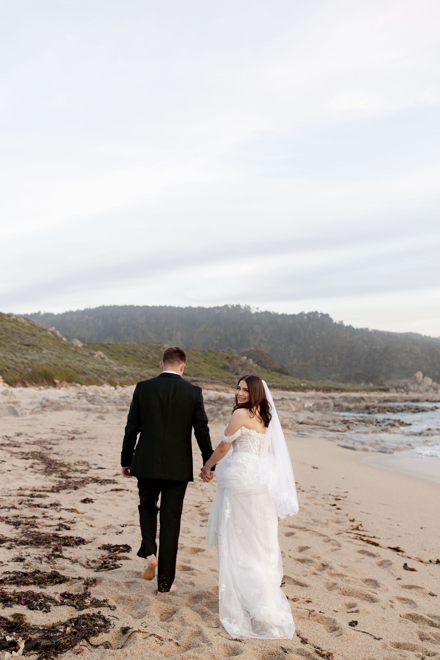 Bride and groom walking hand in hand on a sandy beach, bride glancing back in a white lace gown and veil.