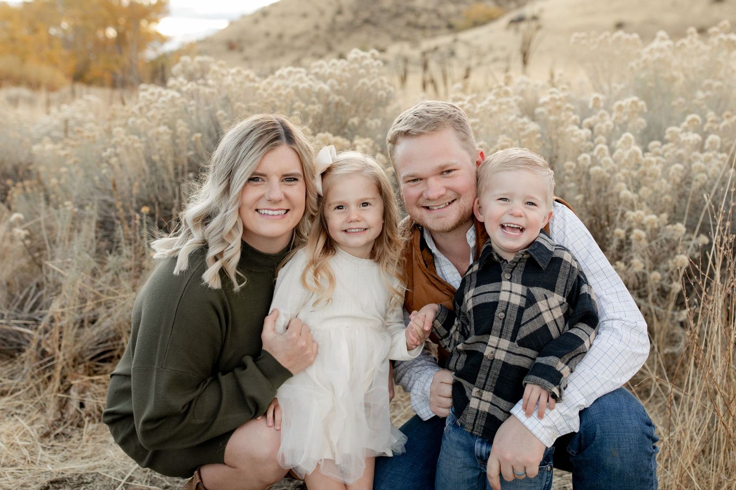 Happy family of four poses outdoors in golden field, parents smiling with young daughter in white dress and toddler son.