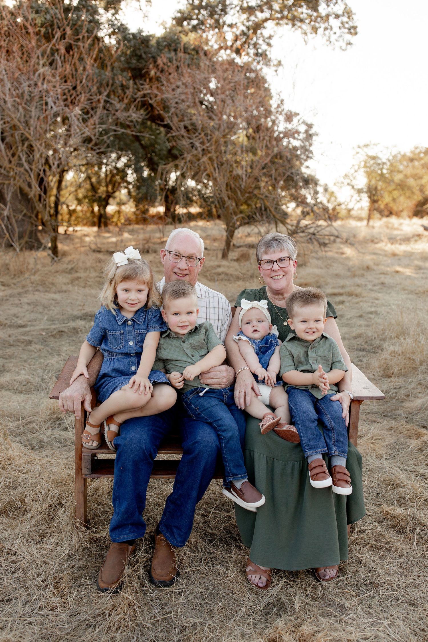 A group sits together on a wooden bench in a rustic outdoor setting with tall grass and trees in the background.
