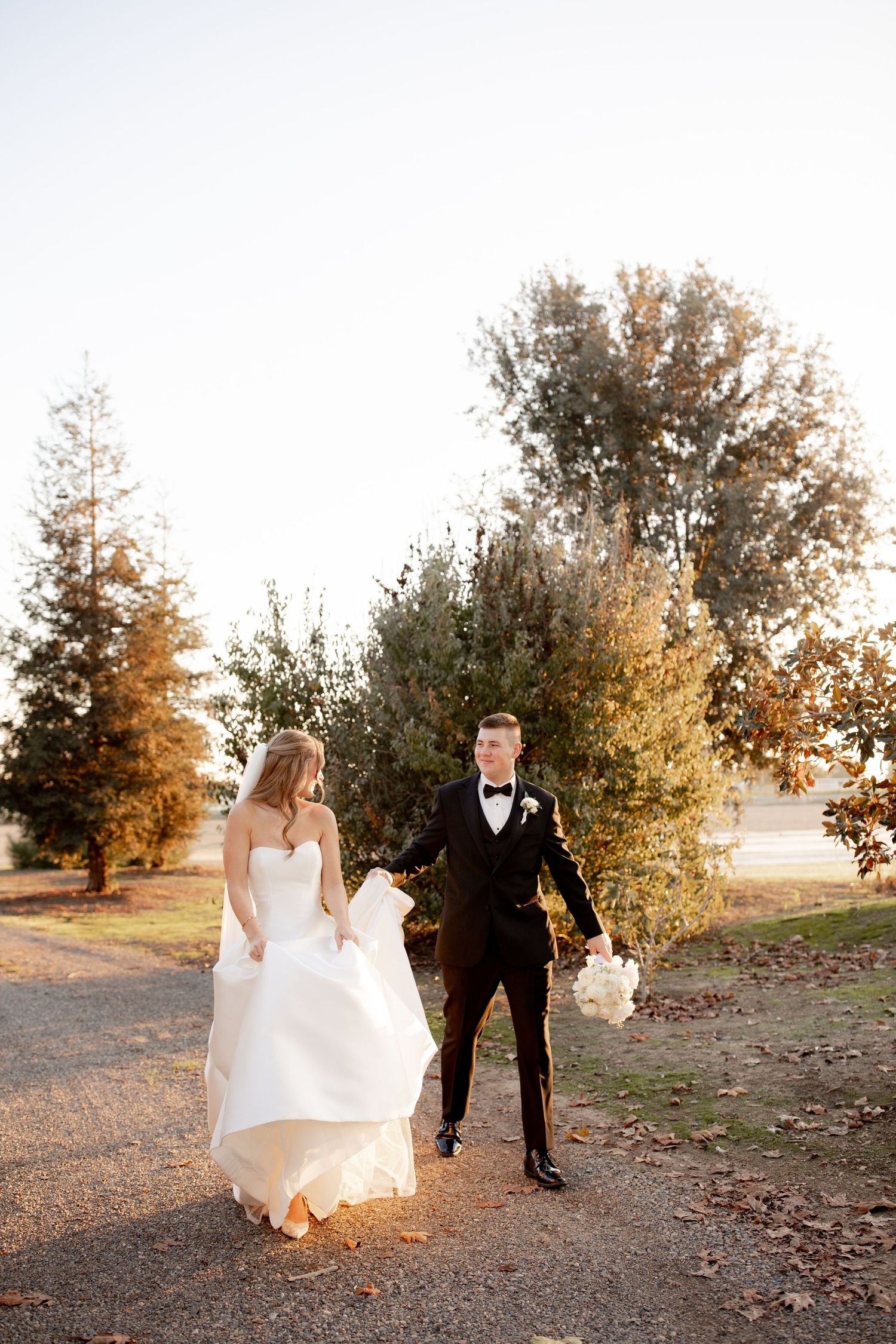 A couple in wedding attire walks along a scenic path during golden hour with trees in the background.