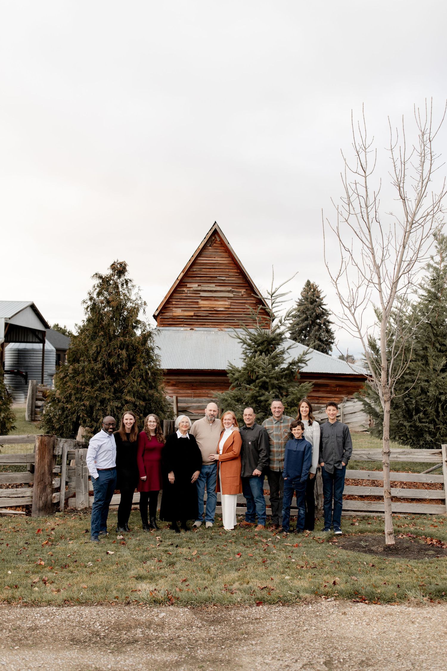 A group of people stand together in front of rustic wooden buildings and bare trees in a rural outdoor setting.
