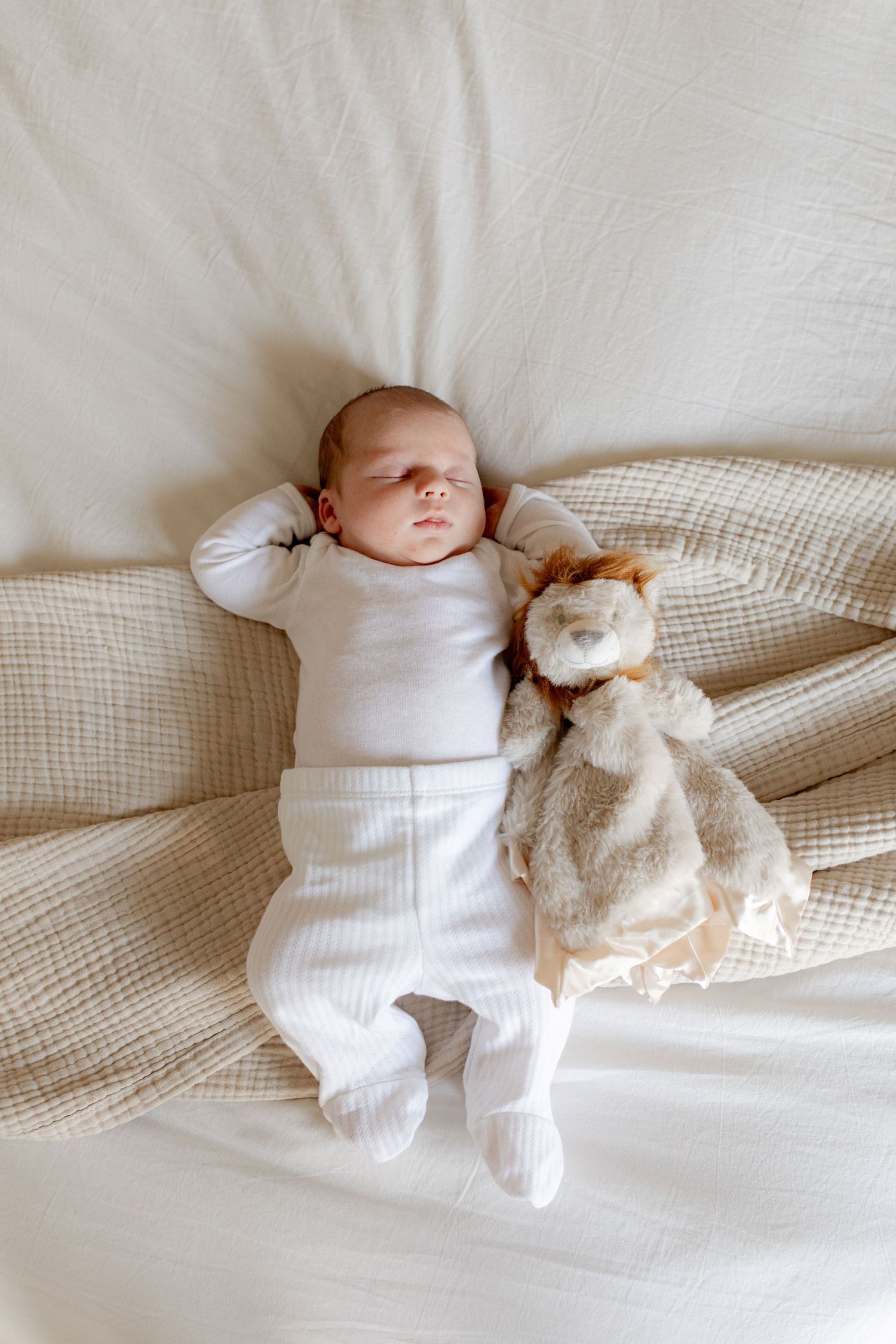 A sleeping baby in white clothing lies peacefully on cream-colored bedding next to a plush bunny toy.