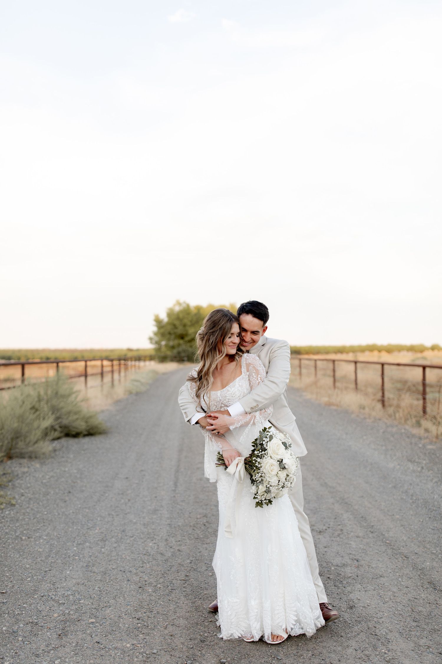 A romantic couple embraces on a dirt road surrounded by rural farmland at sunset, with the bride holding a white bouquet.