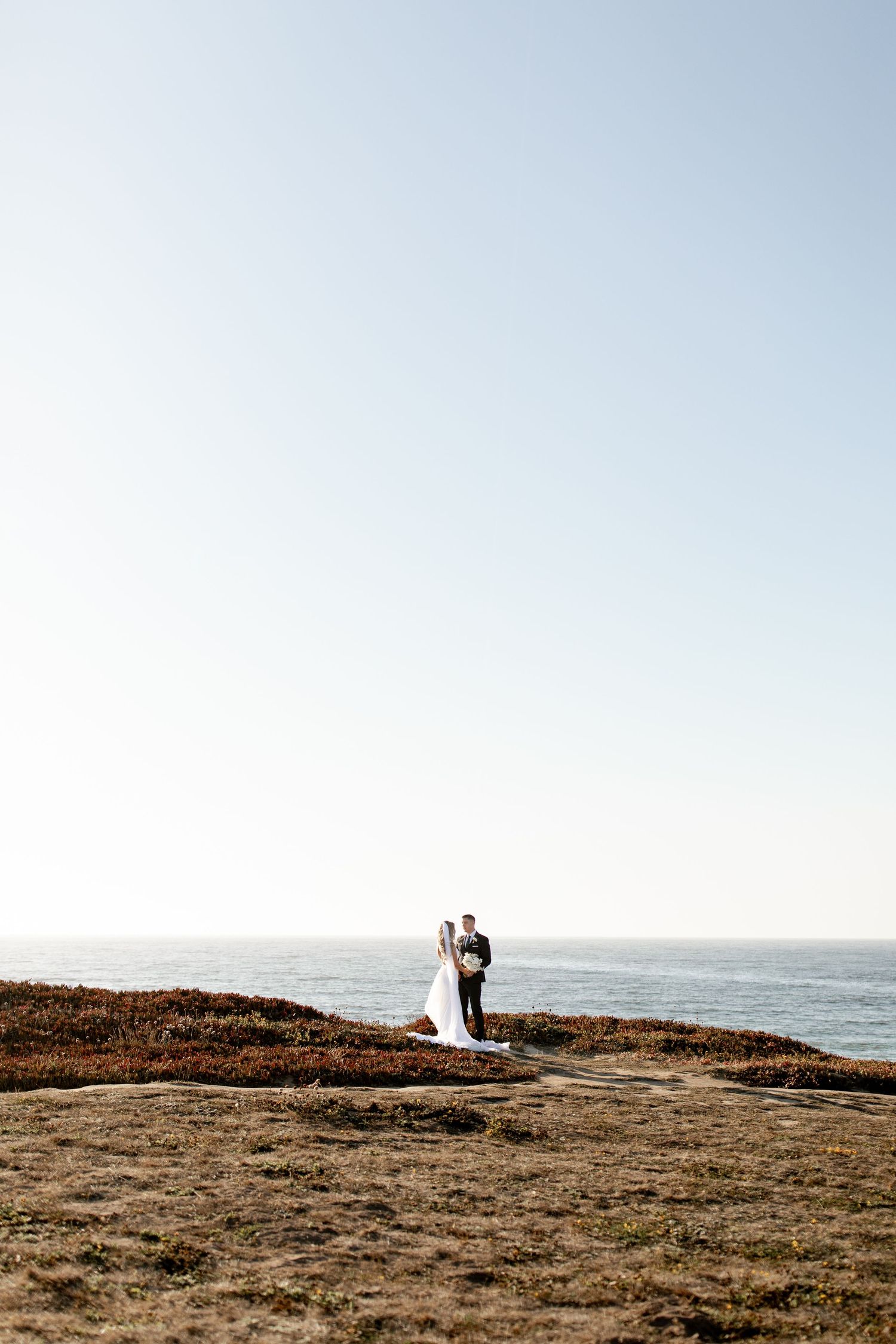A couple stands together on a coastal cliff overlooking the ocean during a romantic photoshoot at sunset.