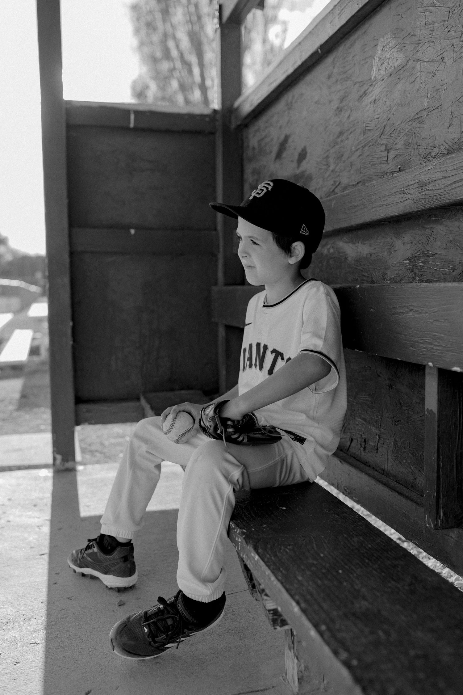 A young baseball player in a Giants uniform sits alone in the dugout during a game.