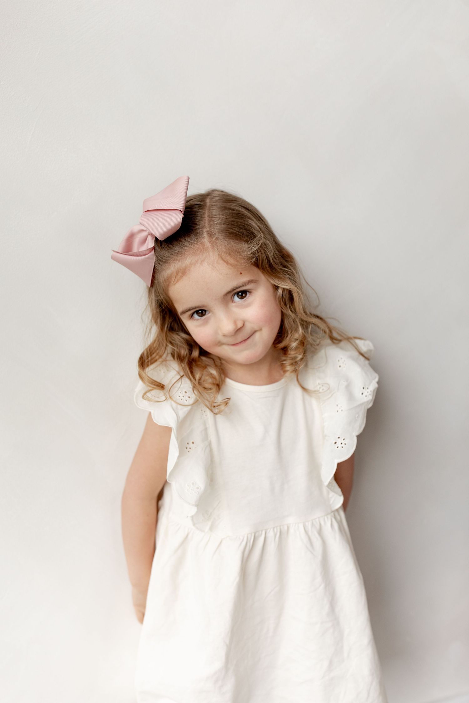Sweet portrait of a child wearing a white eyelet dress and pink bow against a light background.