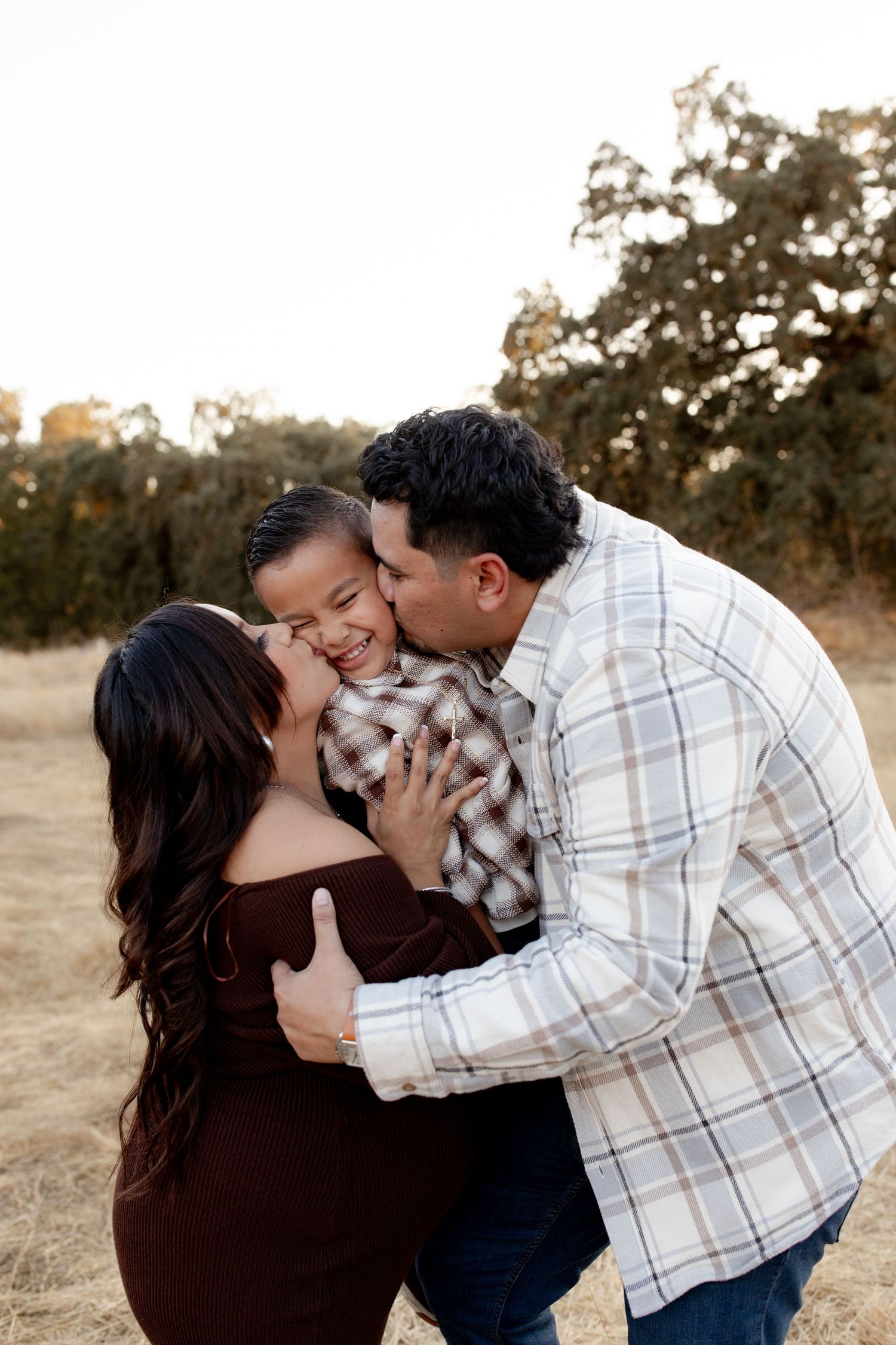 A loving family of three shares a joyful moment outdoors, with parents kissing their laughing young son in a field.