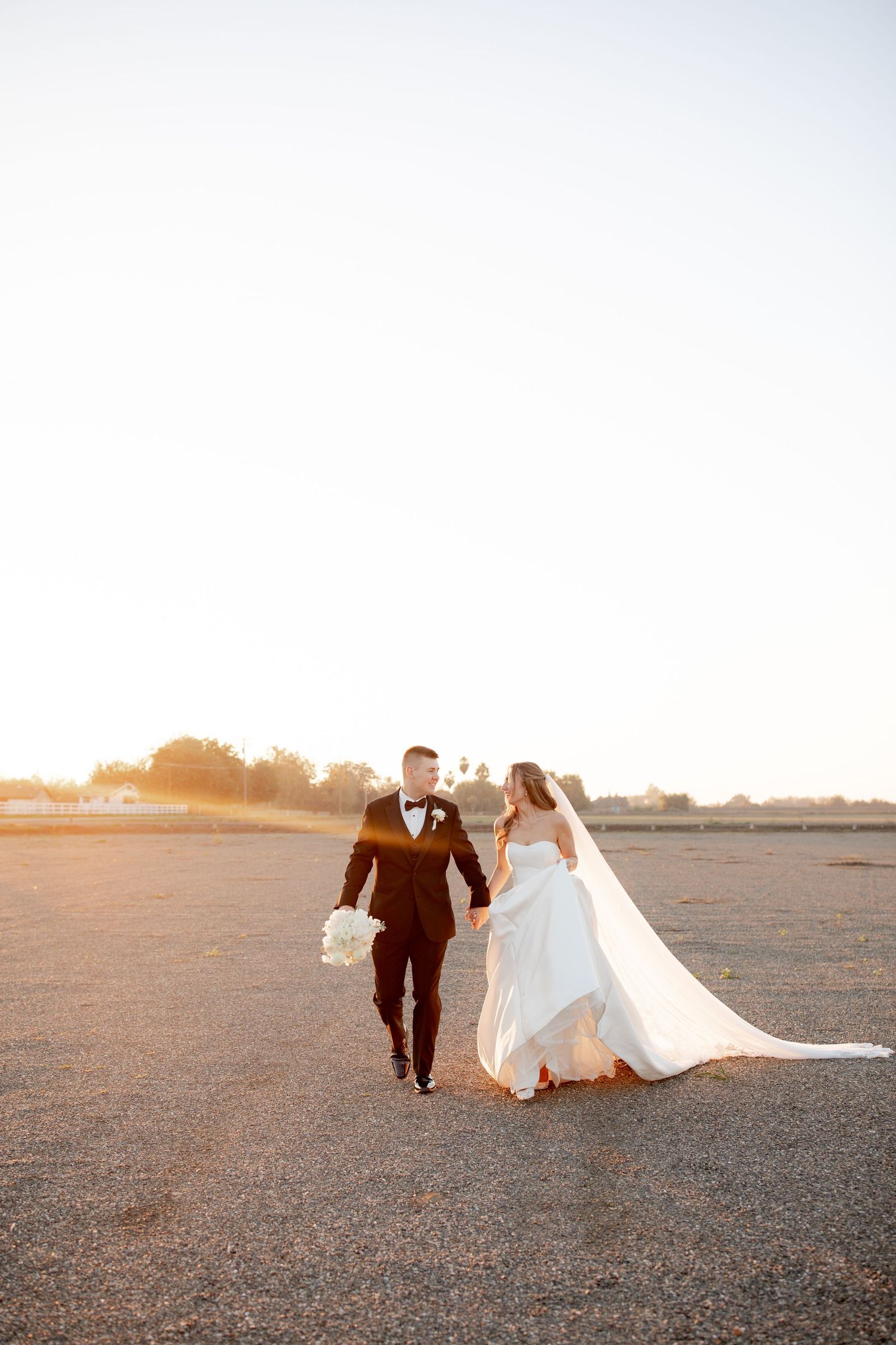 A couple walks together on a paved surface during golden hour sunset, with the bride's white dress and veil flowing behind them.