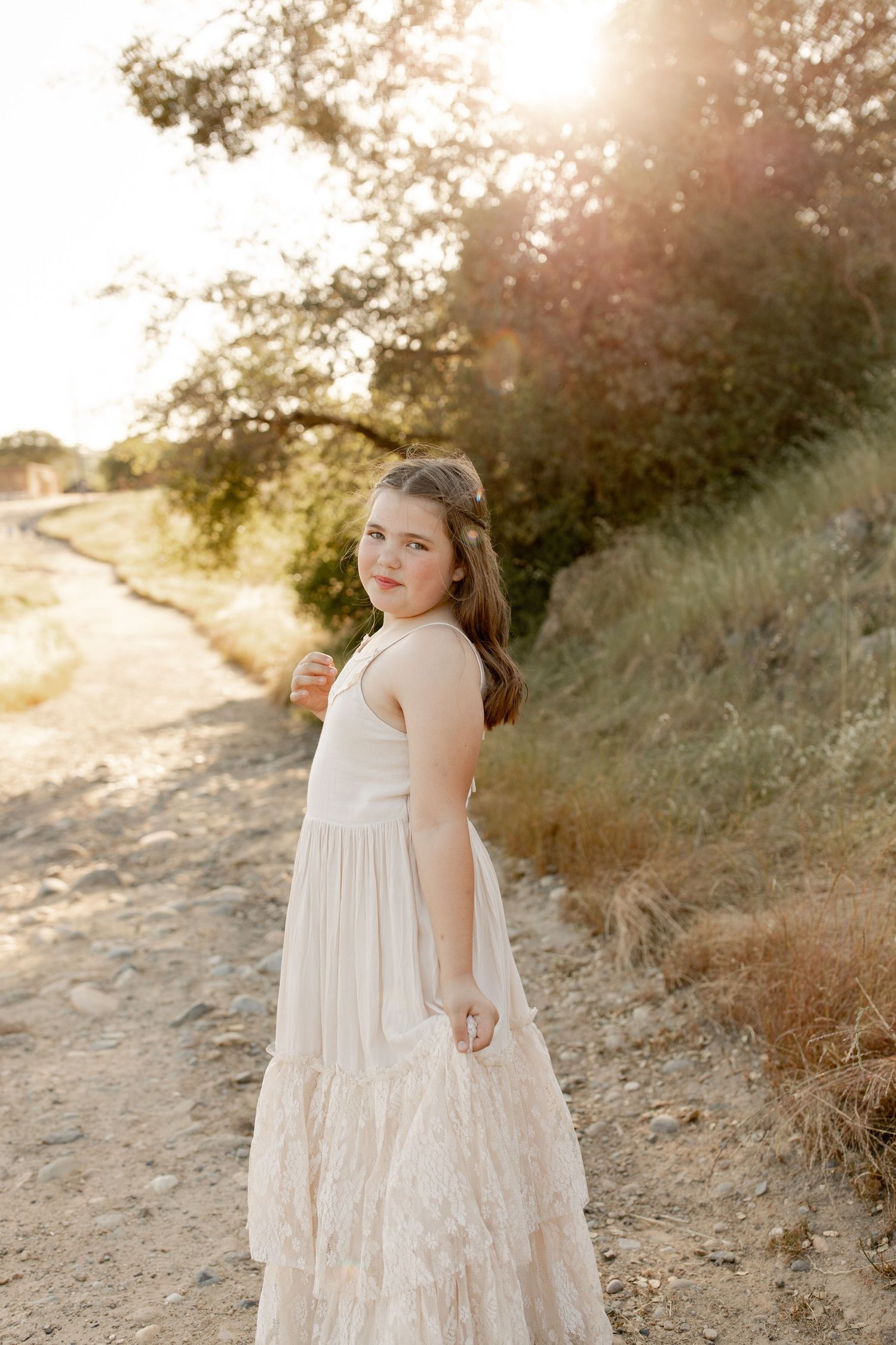 A person in a flowing white tiered dress poses on a sunlit hillside path during golden hour.