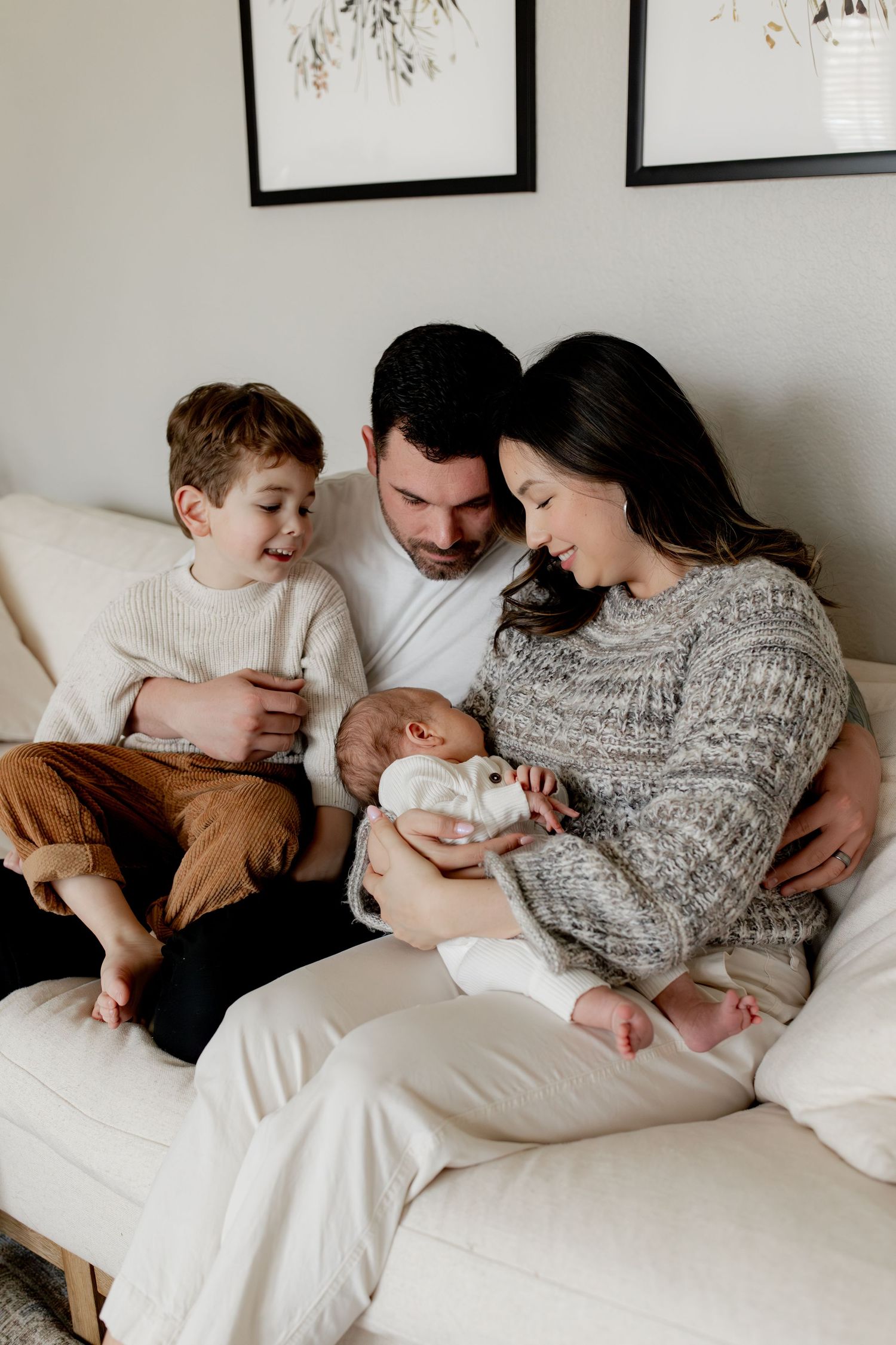 A family cuddles together on a white bed wearing cozy sweaters and casual clothing in a minimalist bedroom setting.