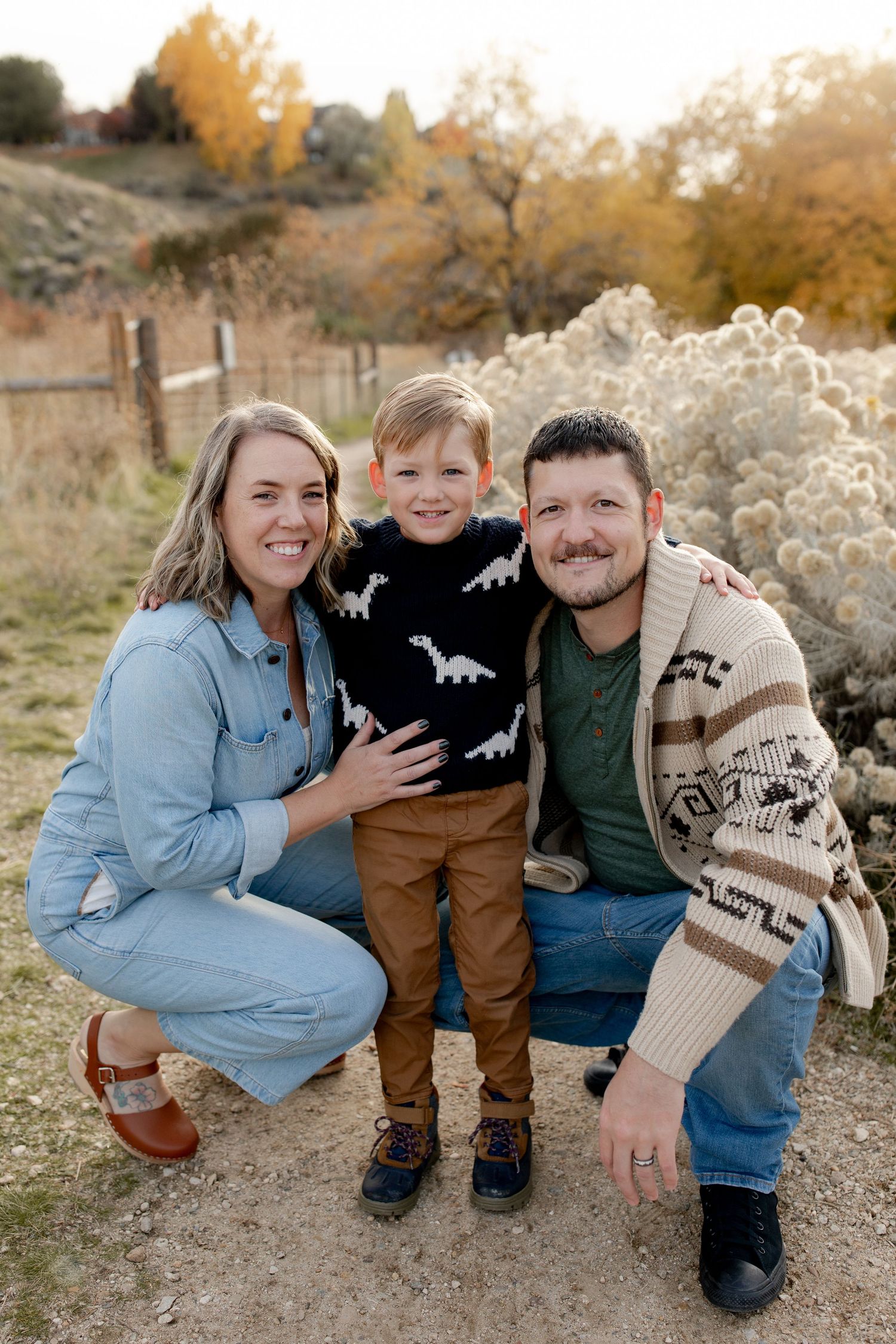 Family of three posing outdoors in autumn, parents crouching beside young boy in dinosaur sweater with fall foliage behind.