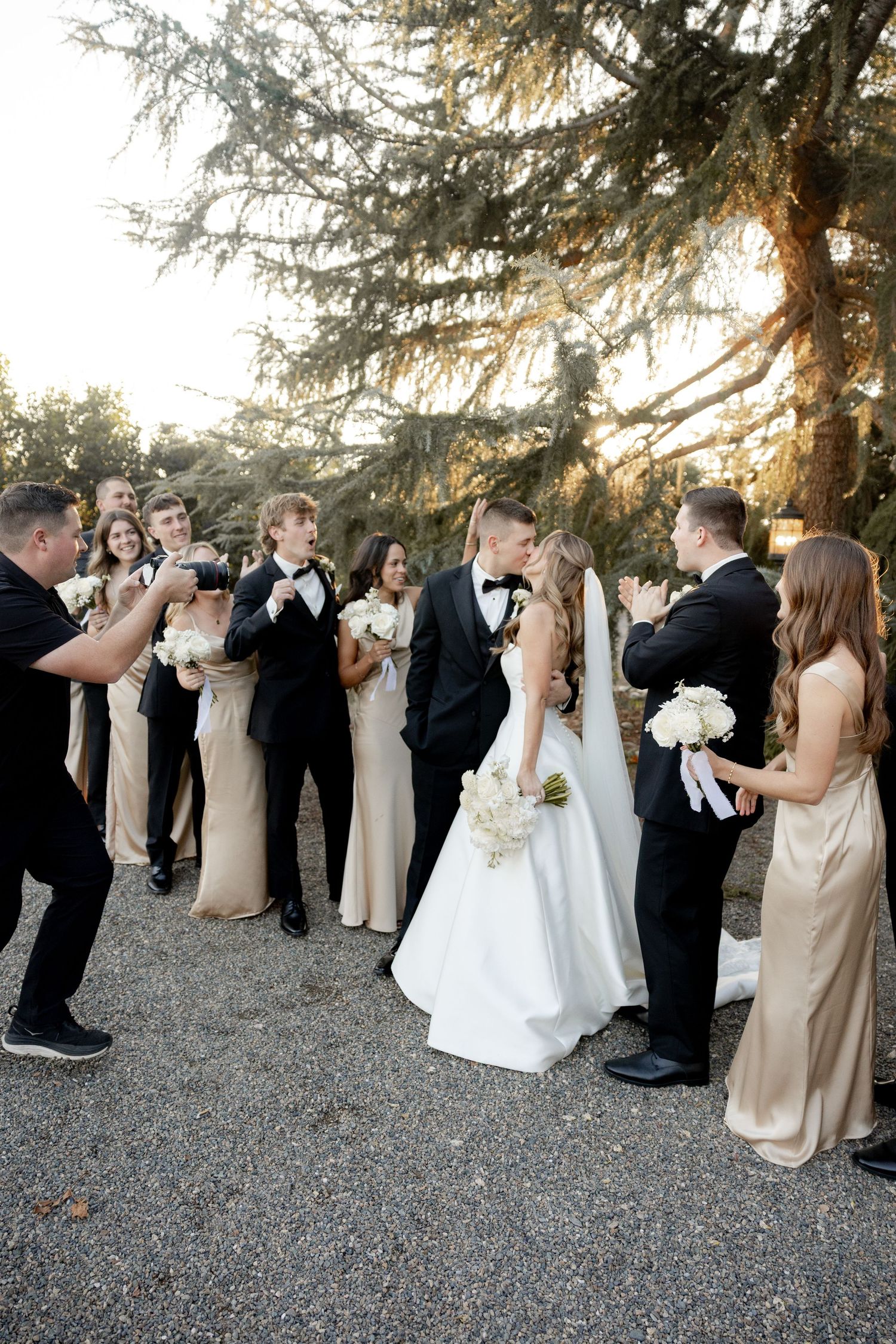 Wedding party gathered outdoors under evergreen trees at sunset during outdoor celebration ceremony.