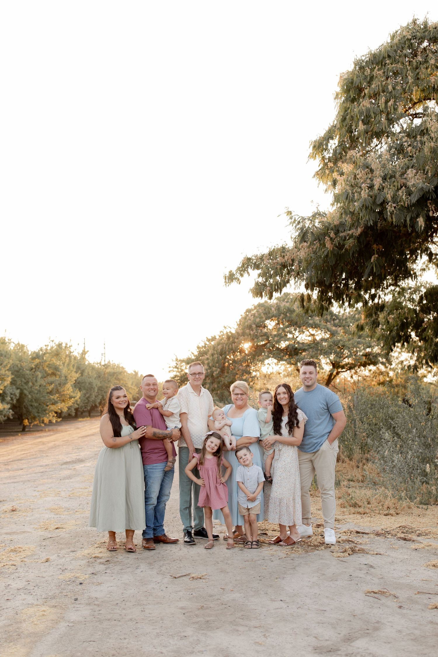 Multi-generational family portrait outdoors on a dirt path, dressed in coordinated pastel outfits, surrounded by trees at golden hour.