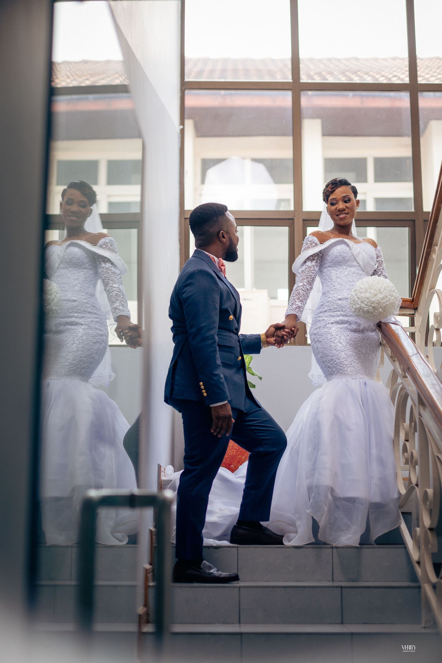 A man and a woman are walking down a staircase, holding hands. The man is wearing a blue suit, and the woman is in a white wedding dress,
