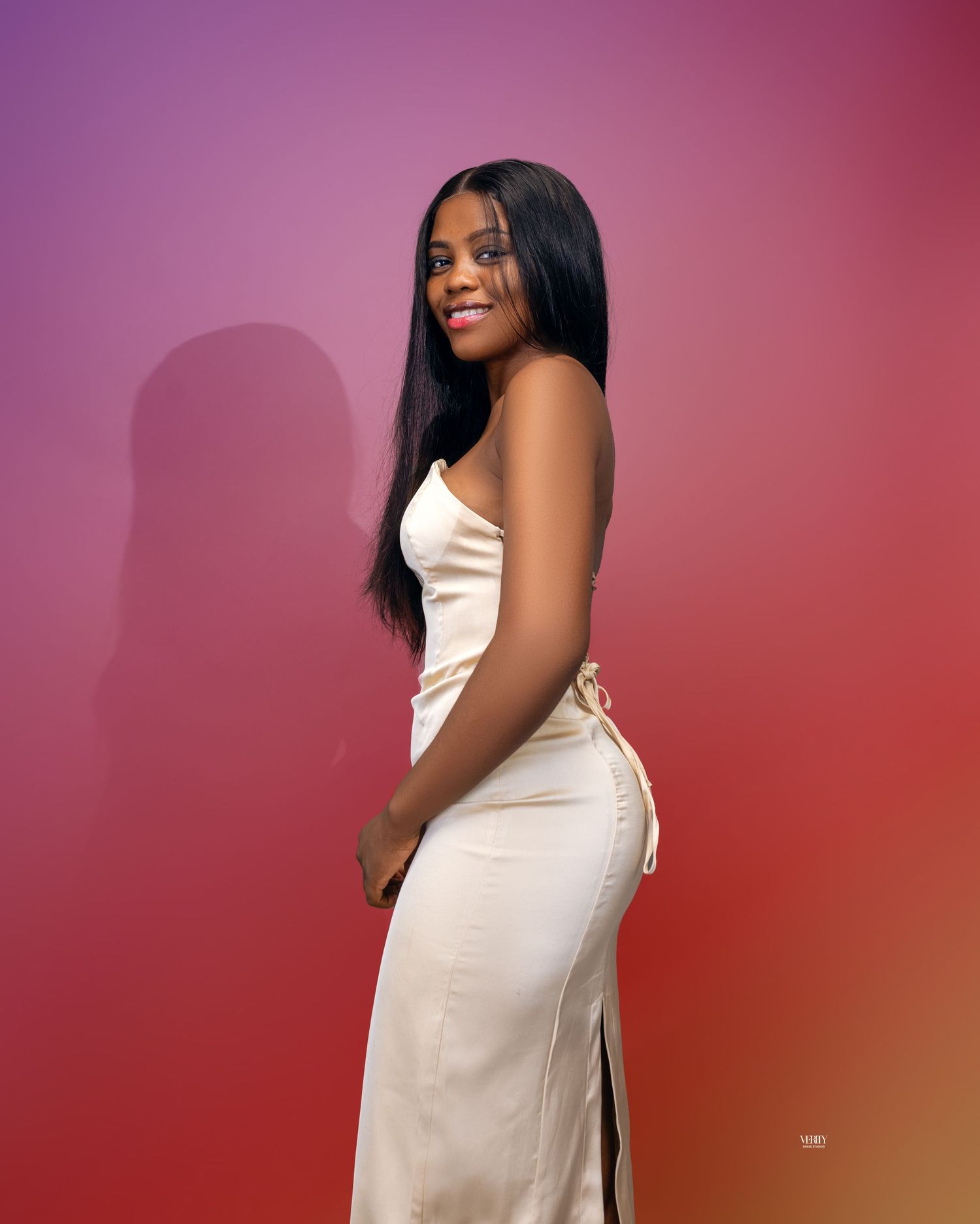 A woman is standing against a reddish looking background and she is wearing an off-white off shoulder dress