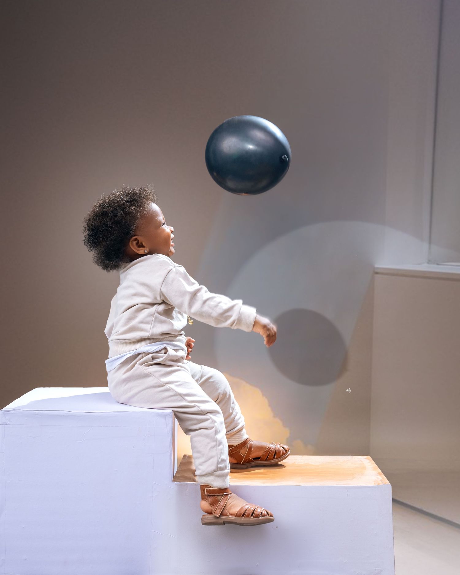 A baby is hitting a balloon and sitting on a white box