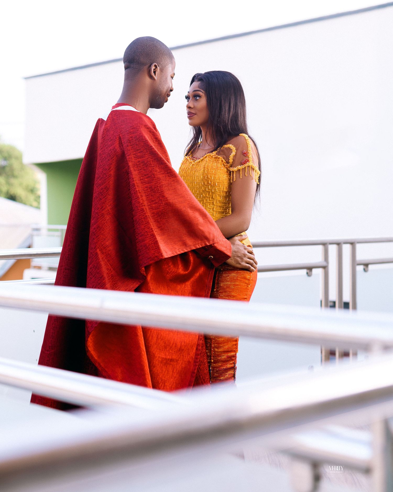 A man and a woman are standing close together on a balcony with a modern railing. The man is wearing a red traditional outfit, and the woman