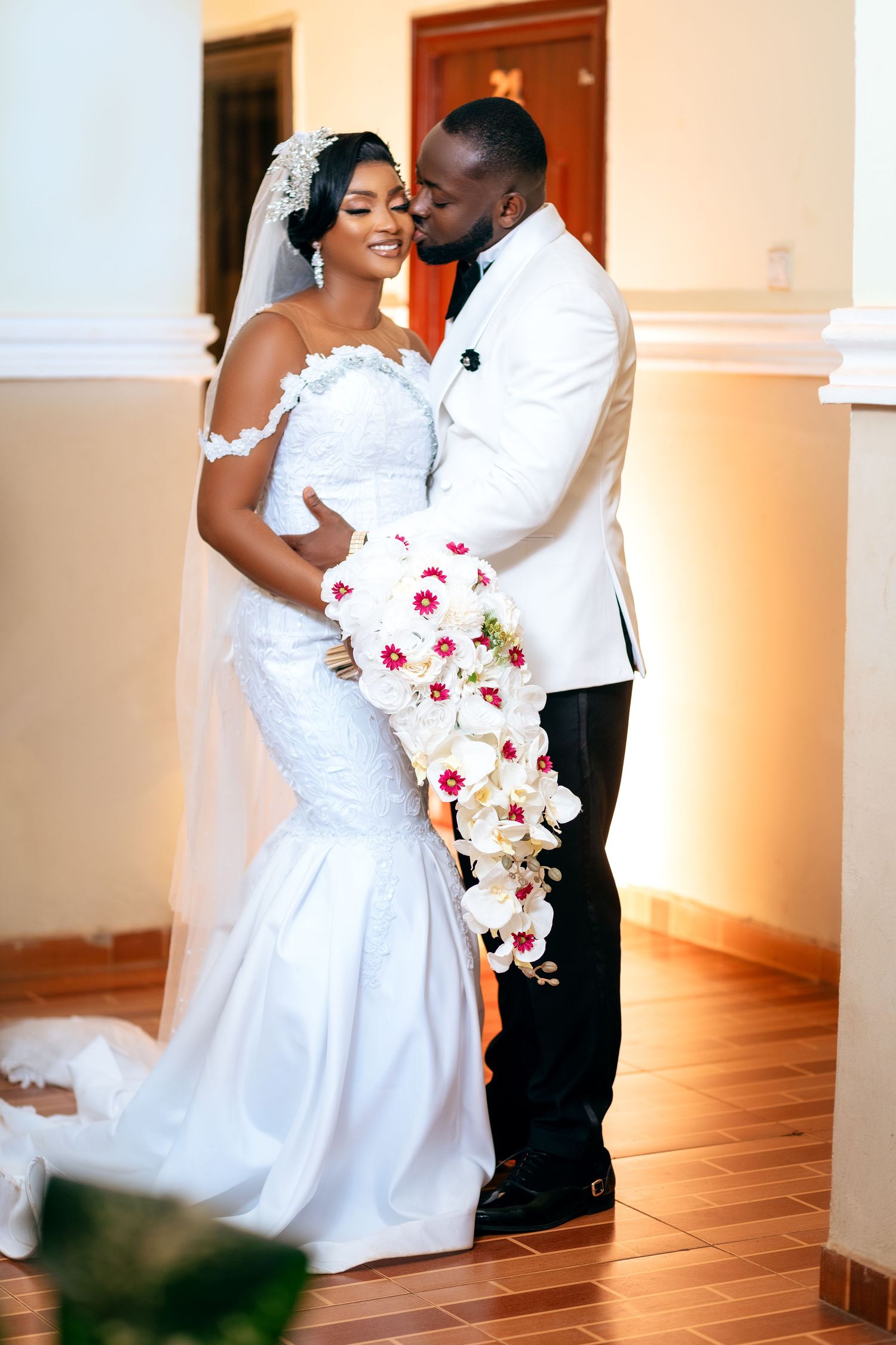 A bride and groom are standing together in an indoor setting. The bride is wearing a white wedding gown with lace details
