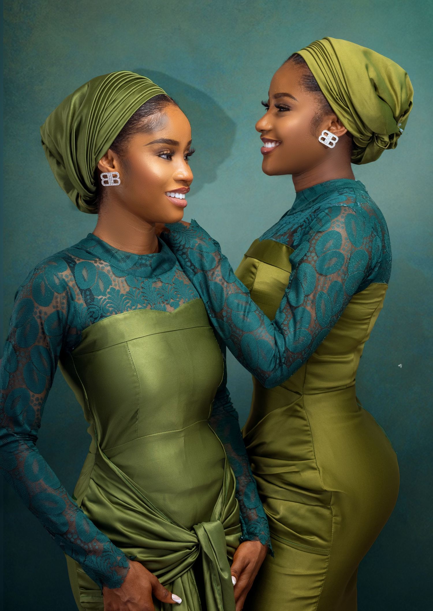 Two women are standing close together, both wearing matching green outfits with intricate lace sleeves and headwraps. One woman has her hand