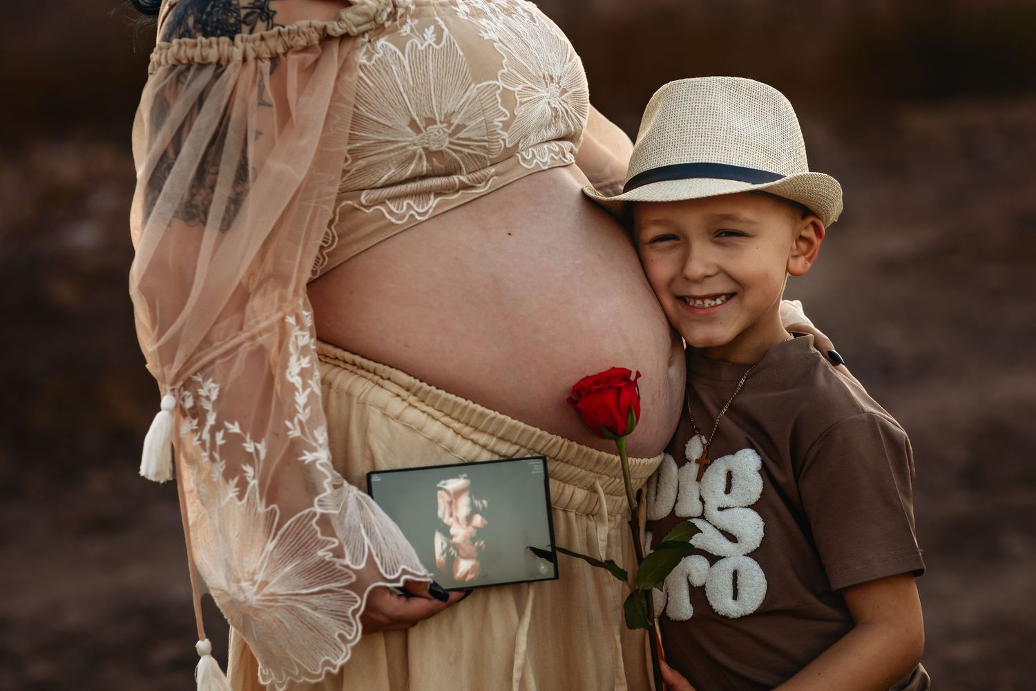 A young child wearing a brown t-shirt with a floral design hugs a woman's pregnant belly adorned with a red flower.